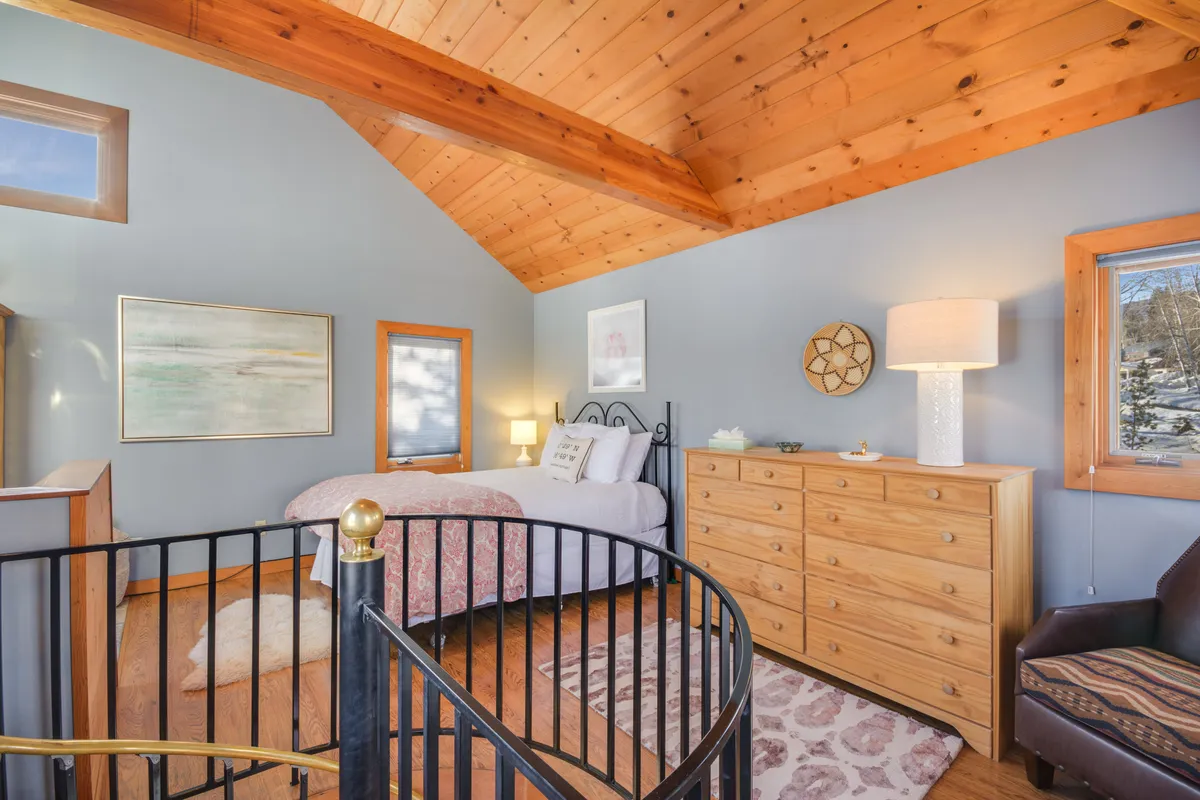 This image showcases a serene bedroom featuring a cozy bed, a dresser, an armchair, decorative artwork, and the distinctive spiral staircase ascending to the upper level. Natural light streams in, creating a peaceful atmosphere ideal for relaxation and quiet moments.