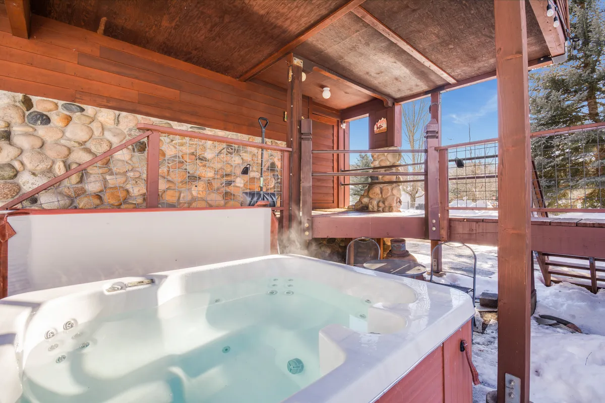 This image showcases a private hot tub set on a wooden deck, bordered by a stone wall and surrounded by snowy scenery. People are seen relaxing in the hot tub and gathering with friends, all while enjoying tranquil views of the winter landscape.