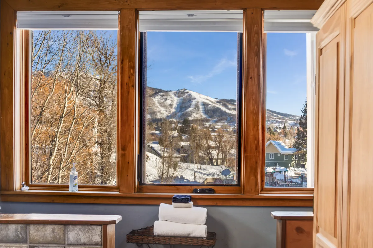 A tranquil bathroom retreat with large windows offering mountain views, neatly arranged towels, and elegant wooden cabinetry. The mood is peaceful, with the option to relax in a bath while enjoying the scenery outside.