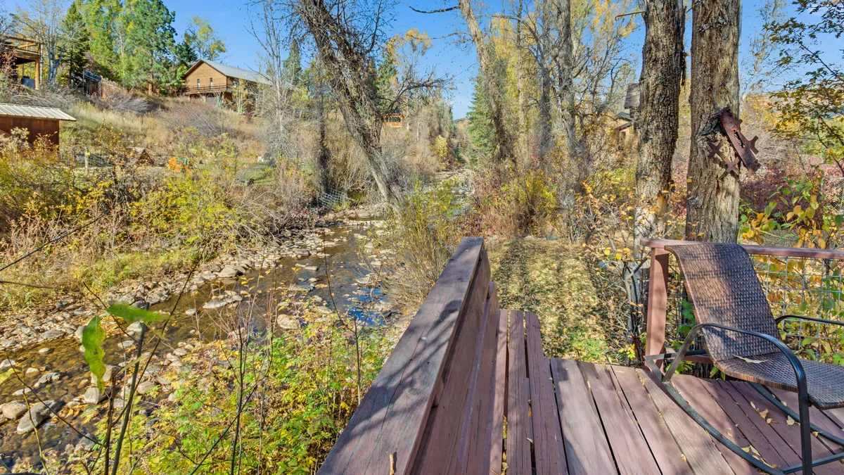 A peaceful creek meanders beside a wooden deck, which is framed by trees and mountain cabins in the distance. The deck offers seating for relaxing by the water, outdoor dining, and bird watching, set against the tranquil backdrop of nature.