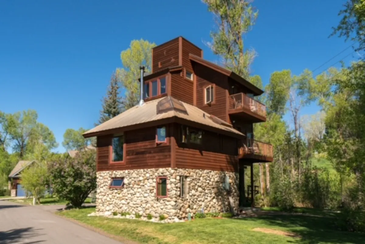 A wide shot showcasing the house’s unique architecture, with a stone foundation, wooden exterior, and balcony railing. The setting blends into the natural landscape, inviting gatherings or quiet moments on the balcony as dusk approaches.