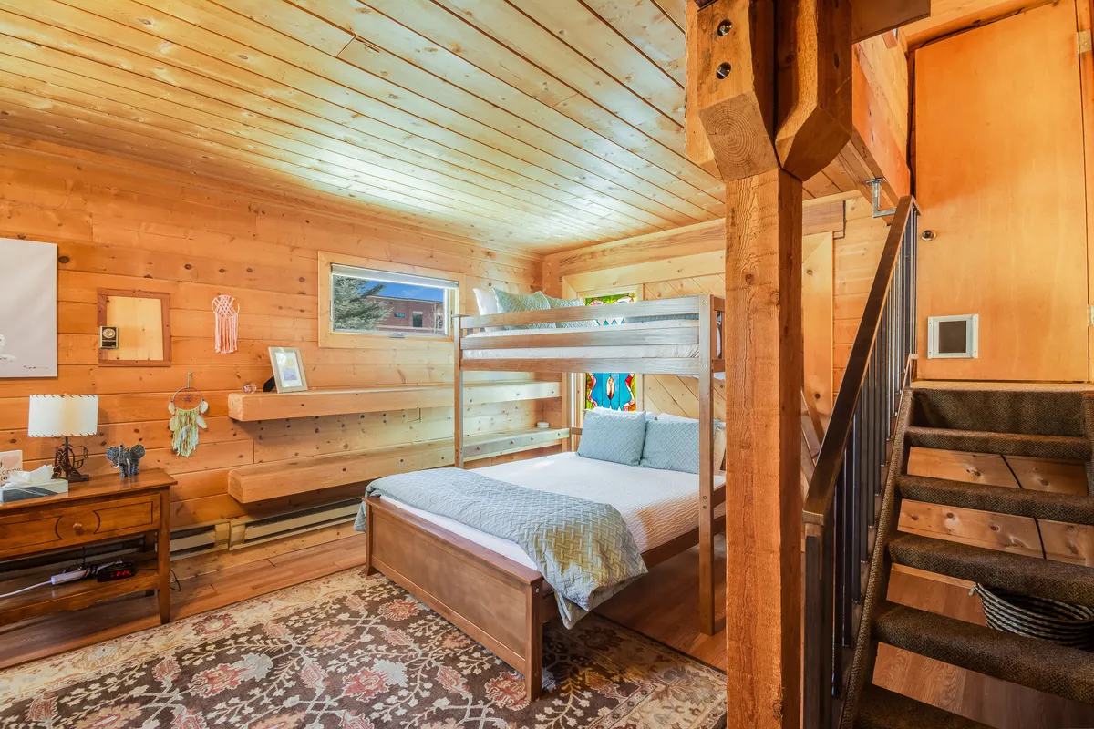 A warm and inviting bedroom with a full-over-queen bunk bed, decorative rug, wooden walls, and a charming stained glass window. The scene is welcoming and ideal for families or groups, reflecting a space designed for both connection and independence.