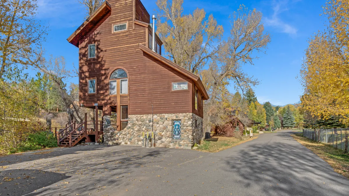 The exterior of a tall wooden structure with a stone foundation, nestled among trees, with a quiet street inviting leisurely strolls or bike rides. The setting appears peaceful and family-friendly, ideal for evening walks or time spent outdoors.