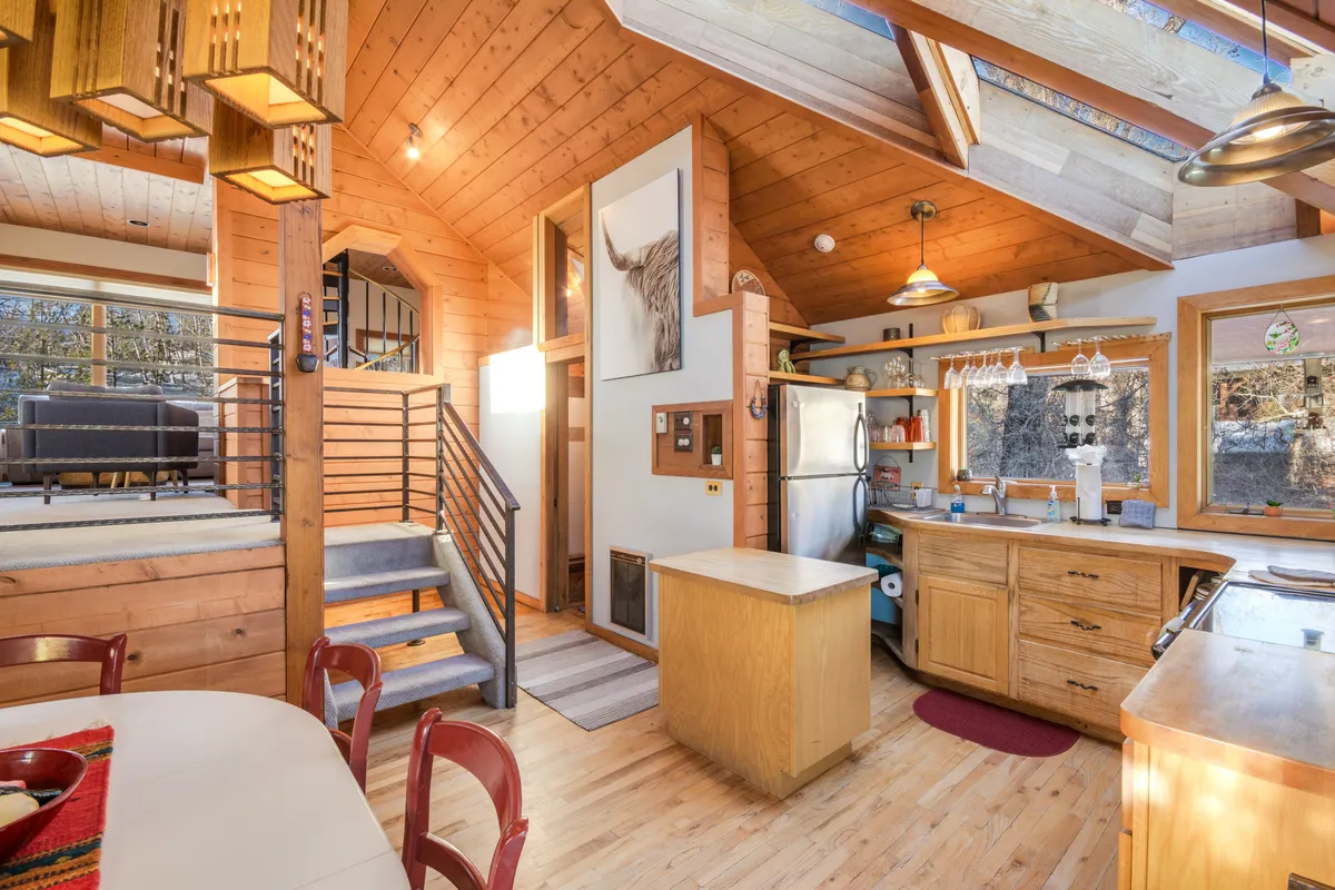 A welcoming, sunlit living space with a prominent staircase, kitchen, large windows, and a dining table. The kitchen skylight filters daylight into an open-plan area that encourages socializing, cooking, and dining together. Plush seating invites relaxation and conversation after a day out.