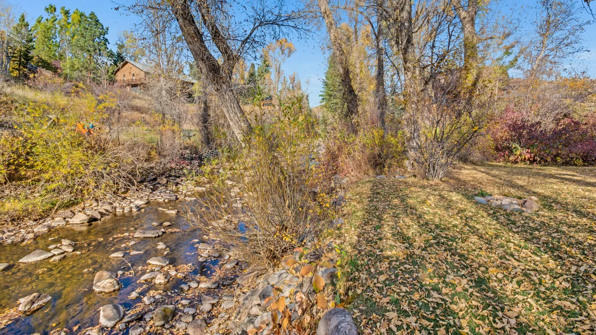 A serene stream bordered by trees and seasonal foliage, with a rocky bank, evokes the picturesque natural setting just steps from the home. The tranquil waterway connects to the idea of a private hot tub overlooking the creek and the proximity to outdoor adventure.