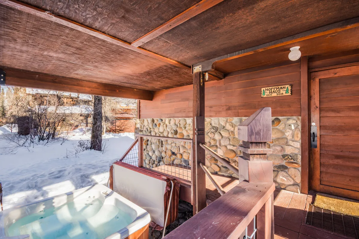 A covered outdoor hot tub set beside the rustic wooden house, with a stone wall and winter scenery visible. The inviting hot tub steams in the cold air, promising relaxation under the stars regardless of season.