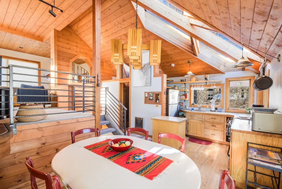 The image showcases a warm, inviting dining area beneath rich wooden ceilings and striking skylights that bathe the room in natural light. A beautiful kitchen is visible, perfect for cooking and gathering, while decorative lighting and thoughtful décor create a cozy, welcoming atmosphere ideal for family meals or game nights.