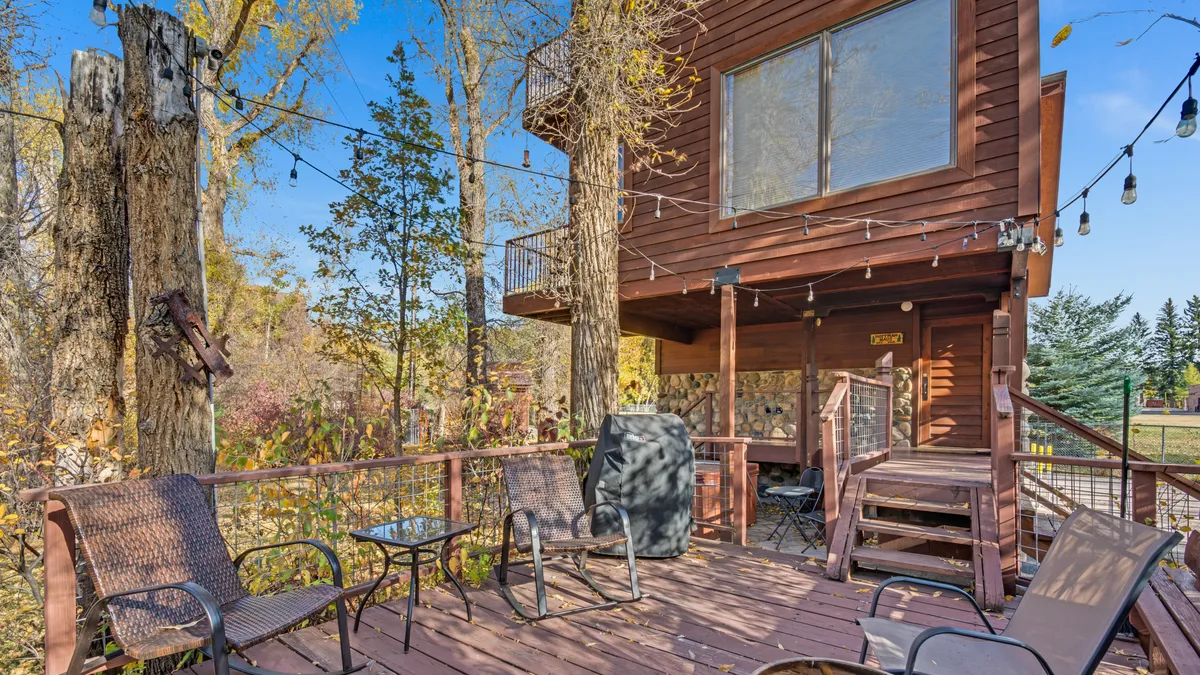 A cozy deck seating area adorned with string lights sits outside the house, inviting guests to relax, entertain, or simply enjoy the view through large house windows. The setting hints at both daytime and nighttime enjoyment, surrounded by trees and nature.